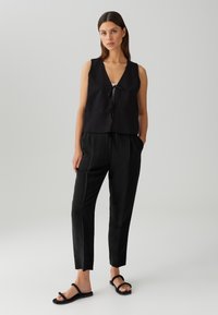 Gilet sans manches noir avec un col en V et un détail de nœud, associé à un pantalon noir effilé et des sandales plates à brides, sur un fond clair.