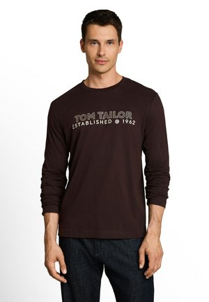 Man wearing a dark long-sleeve shirt with "Tom Tailor Established @ 1962" text, paired with dark jeans, standing against a white background.