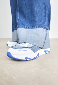 Modré a bílé tenisky Karl Lagerfeld nošené s ohrnutými dvoubarevnými modrými džíny na světlém podkladě.