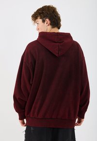 Burgundi színű, oversized kapucnis pulóver texturált felülettel, zsinóros kapucnival és elasztikus derékkal. A modell hátulról látható.