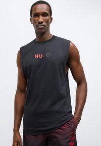 Črna brezrokavnik iz bombaža z okroglim izrezom; na prsih je logotip "HUGO" v rdeči in sivi barvi.