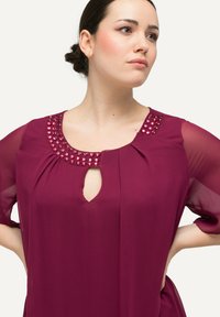 Blouse mauve avec un col rond orné de clous roses brillants. Les manches transparentes et une découpe en forme de œil au front ajoutent des détails de design.