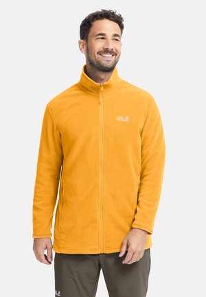 Man glimlacht en draagt een felgele fleecejack met rits, voorzakken en het Jack Wolfskin-logo op de borst.