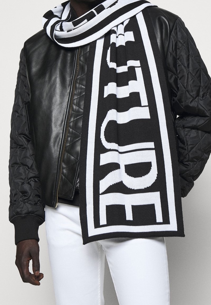 Homme portant une veste en cuir matelassée noire, un pantalon blanc et une écharpe rayée noire et blanche avec un grand texte enroulé autour du cou.