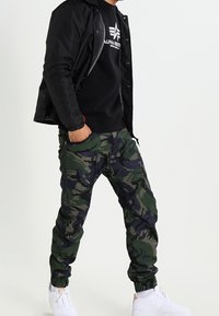 Kamouflage joggers med gröna och svarta mönster, elastiska ärmslut och sidofickor, i kombination med en svart sweatshirt och en svart jacka. Vita sneakers.