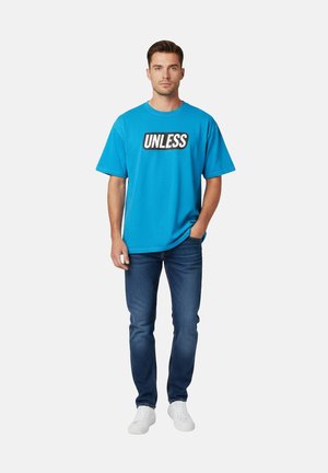 Jeune homme debout portant un t-shirt bleu avec le texte "UNLESS", un jean bleu foncé, des baskets blanches, une main dans la poche.