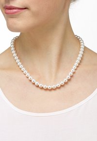 Weiße Perlenkette mit glatten, runden Perlen, die zusammen aufgezogen sind und am Hals gegen ein helles Oberteil liegen.