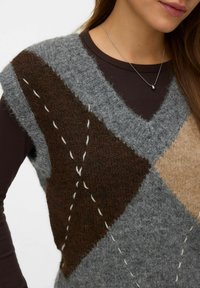 Chaleco de punto en tonos gris y marrón con un patrón de diamantes, adornado con puntadas contrastantes y un acabado texturizado. Combinado con una camisa oscura.