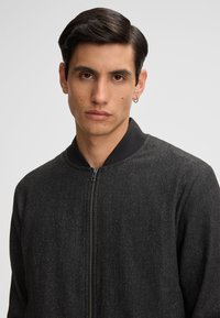 Giacca bomber nera con finitura testurizzata, dotata di chiusura con zip e colletto a coste in contrasto. Vestibilità tailored, design minimalista.