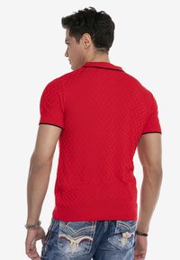 Rotes Polohemd mit kurzen Ärmeln und strukturiertem Zickzackmuster, ausgestattet mit einem schwarzen Kragen und gerippten Bündchen, kombiniert mit blauen Jeans.