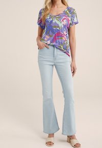 Nainen, jolla on päällä vaaleansiniset leveälahkeiset farkut, beige korkokenkä, ja lyhythihainen kukallinen violetti paita, seisoo käsi taskussa.