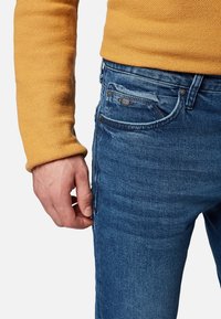 Jeans de denim azul con un corte ajustado, que presentan un diseño clásico de cinco bolsillos, costuras en contraste y una manga de suéter de punto mostaza texturizado visible.