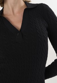 Pull noir en maille avec un motif en cable, présentant un col et un bouton unique. La texture est douce et côtelée.