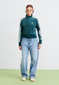 Casaco de veludo verde com fecho éclair e riscas brancas, combinado com jeans de perna larga azul claro e ténis brancos sobre um chão verde claro.