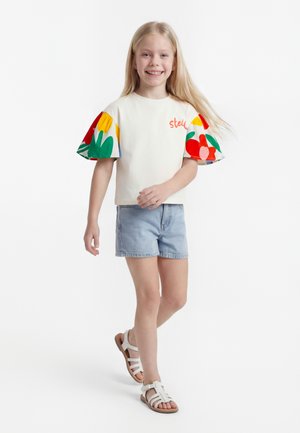 Κοπέλα που χαμογελά με μακριά ξανθά μαλλιά που περπατάει, φοράει λευκό τοπ με πολύχρωμα φλοράλ φουσκωτά μανίκια, τζιν σορτς και λευκά σανδάλια.