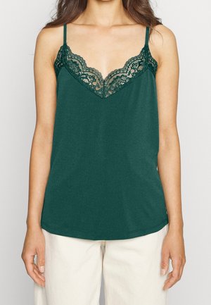 Kobieta w ciemnozielonej bezrękawnikowej bluzce z koronkowym wykończeniem przy dekolcie oraz białych spodniach, stojąca na tle jednolitym.