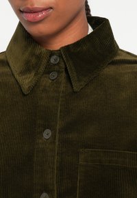 Mørkegrøn corduroy skjorte med krave, der har en knaplukning foran og en stiklomme. Teksturen er ribbet, og knapperne er mørke med en subtil glans.