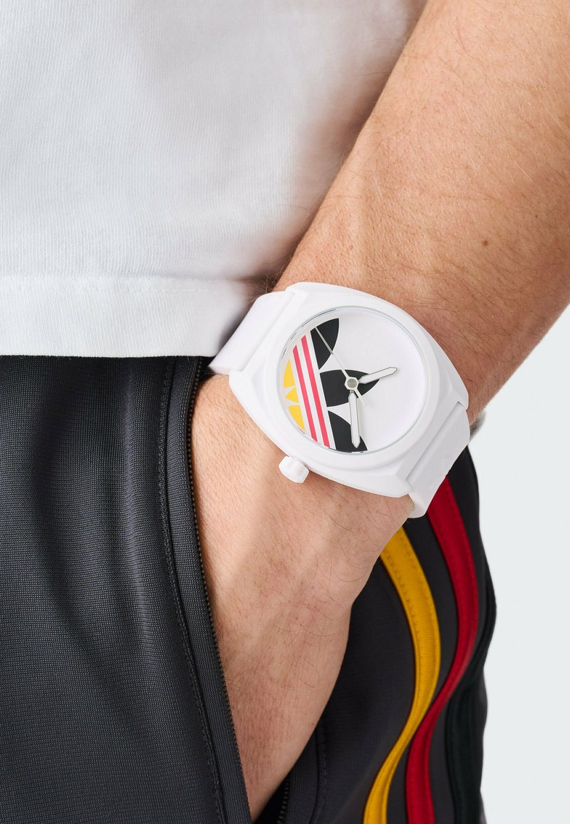 Reloj blanco con esfera redonda, que presenta un diseño gráfico en negro, amarillo y rosa. Fabricado con una correa flexible, se lleva en la muñeca.