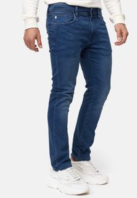 Jeans in denim blu con vestibilità slim, caratterizzati da cuciture a contrasto e design a cinque tasche. Indossati con scarpe sportive bianche.