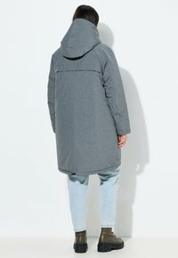 Chaqueta acolchada gris de longitud media con capucha, que presenta una tela texturizada y un dobladillo ligeramente redondeado. Lleva puesta sobre un denim claro y botas.