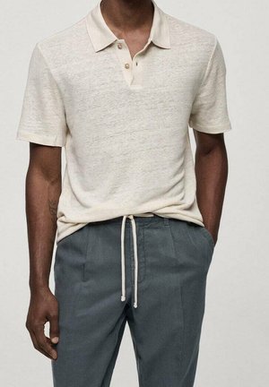 Mann trägt ein beiges Polohemd mit kurzen Ärmeln, in graue Hose mit Tunnelzug gesteckt, steht mit einer Hand in der Tasche.