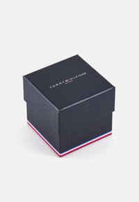 Caja cuadrada negra con tapa, que presenta el logo "TOMMY HILFIGER WATCHES" en blanco en la parte superior y una franja roja, blanca y azul en la base.