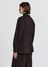 Blazer marrone su misura con una texture liscia, rever a lancia e polsini con bottoni. Indossato sopra un dolcevita scuro e aderente.