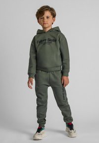 Conjunto de sudadera con capucha y pantalones de chándal en verde oliva con el texto "Future Skate". Tejido suave, puños acanalados y aperturas en los tobillos. Zapatillas con acentos multicolores.