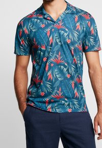 Camisa de manga corta en azul teal con un patrón floral tropical que presenta hojas y flores rojas y verdes. Cuello con solapa, tejido ligero.
