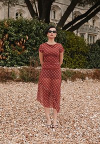 Rochie bordo cu model, mâneci scurte, fustă în straturi, material ușor transparent și detaliu subtil cu volane. Purtată cu ochelari de soare negri și sandale.