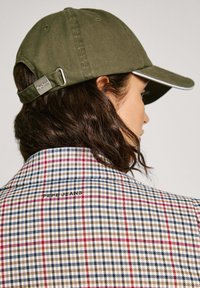 Cappello verde oliva con visiera curva e bordo a contrasto. Indossato con un blazer a quadri in tonalità di rosso, blu e beige.