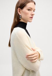 Cardigan in maglia a maniche lunghe di colore crema, morbido e peloso, con scollatura a V, indossato sopra un dolcevita nero. Orecchini rotondi argentati aggiungono un tocco metallico.