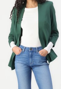 Zelený blejzr s přiléhavým střihem, dlouhými rukávy a hladkým povrchem, v kombinaci s bílou košilí a modrými džíny s vysokým pasem.