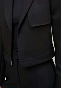 Musta lyhyt blazer herringbone-kankaasta, jossa on leikattu kaulus, etutaskun läppä ja jämäkkä olkapäiden muotoilu.