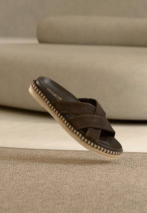 Bruine suède sandaal met gekruiste bandjes, platte zool en geweven jute details. Getoond boven een getextureerd beige oppervlak.