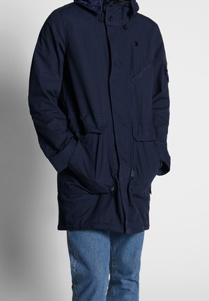 Parka de color azul marino con capucha forrada, botones frontales, dos bolsillos grandes y tela texturizada. Lleva puesta sobre unos vaqueros de color azul claro.