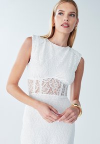 Robe en dentelle blanche avec manches courtes et structurées, présentant une partie centrale en dentelle transparente. Accentuée par un simple bracelet en or au poignet.