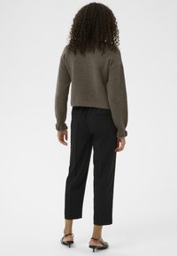 Ženska s kodrasto pričesko, oblečena v rjav obrezan pulover, črne obrezane hlače in črne čevlje na peto s slingback ukrivljenjem, stoji obrnila se proti nas.