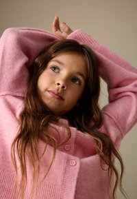 Cardigan rosa chiaro con trama a coste e piccoli bottoni rotondi, indossato sulle braccia, che mostra una vestibilità rilassata e un aspetto accogliente.