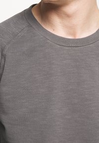 Camisa gris de manga larga con una tela texturizada y ligeramente jaspeada. Presenta un escote tipo crew y un detalle de costura visible en los hombros.
