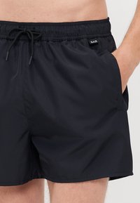Čierne plavkové šortky vyrobené z ľahkej, hladkej tkaniny. Elastický pás s tkacím pásom, bočné vrecká a logopatch značky na prednej strane.