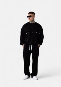 Sweatshirt noir avec texte graphique blanc, coupe décontractée, associé à un jogging noir avec cordons blancs. Des baskets blanches complètent le look.