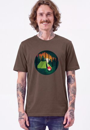 Lachende man met tatoeages die een bruin T-shirt draagt met een kampeerscène met een tent, kampvuur, bomen en zonsondergang.