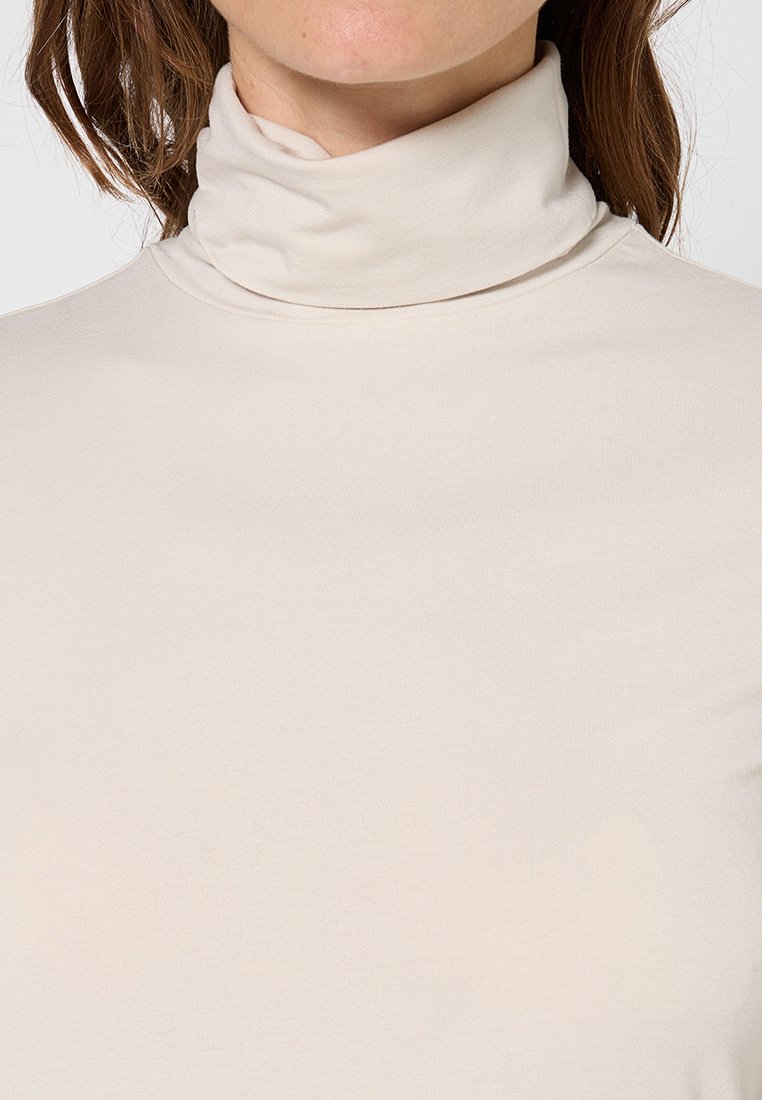 Helles beiges Rollkragenoberteil mit einer sanften, glatten Textur, das über einen umgeschlagenen Kragen und eine enge Passform im Bereich von Hals und Schultern verfügt.