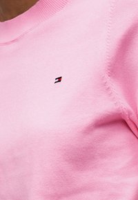 Κοντινή λήψη ενός ατόμου που φοράει ένα ανοιχτό ροζ πουλόβερ με ένα μικρό, οριζόντιο κόκκινο, λευκό και μπλε λογότυπο σημαίας στο στήθος.