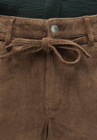 Prunoogid püksid, millel on seotav kangast vöö ja nähtav metallnupp vöökohtas musta särgi peal.