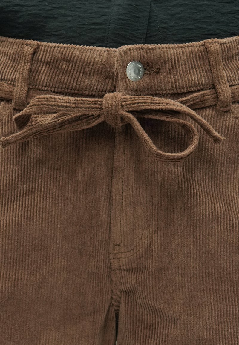 Pantalon en velours côtelé marron avec une ceinture en tissu nouée et un bouton en métal visible à la taille, porté avec une chemise noire.