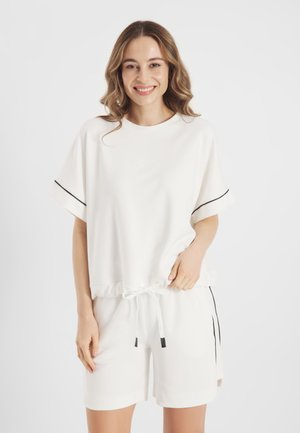 Młoda kobieta uśmiechająca się, ubrana w biały top z krótkimi rękawami z czarnym wykończeniem oraz pasujące krótkie spodenki z paskami po bokach, stojąca na jednolitym tle.