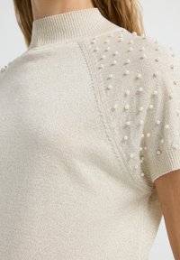 Beige gebreid top met een hoge hals, textuurstof en korte mouwen versierd met kleine witte parelachtige decoraties.