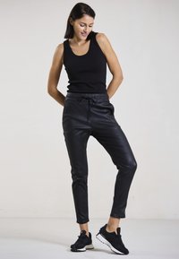 Schwarzes ärmelloses Tank-Top kombiniert mit schwarzen Jogginghosen aus Kunstleder, die über einen elastischen Bund und Seitentaschen verfügen. Schwarze Sportschuhe.